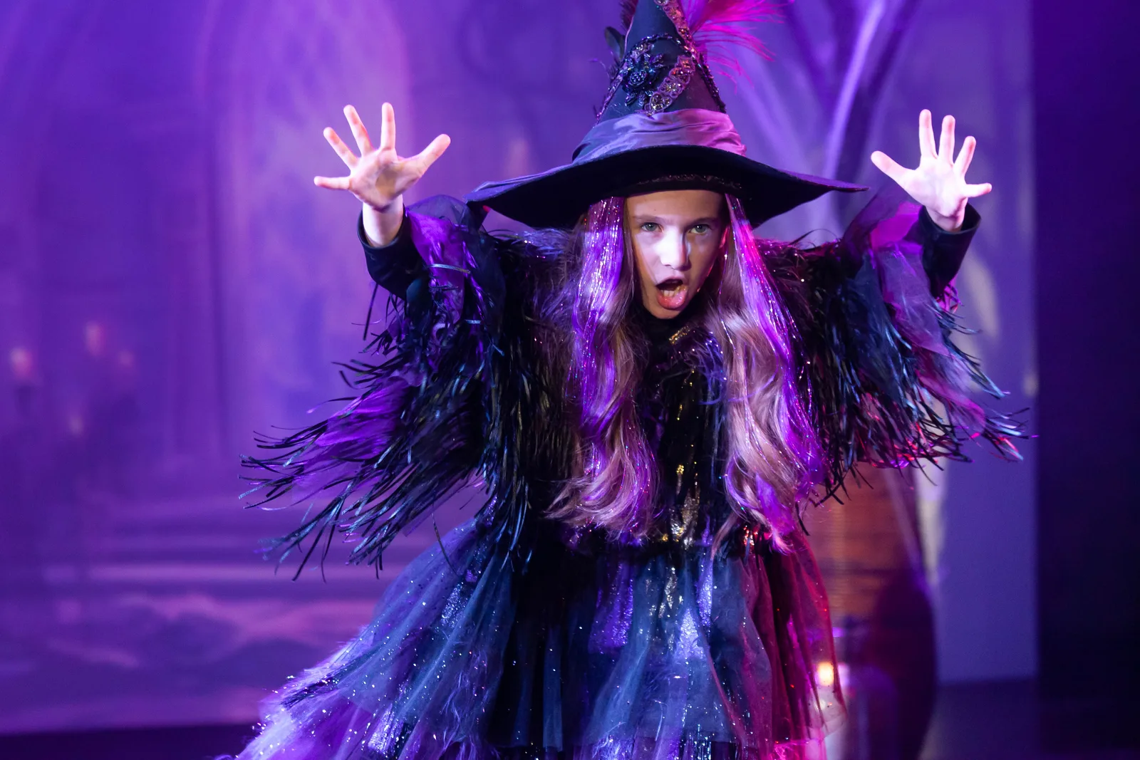 A Starbright student in a feathered witch costume and pointed hat casting a spell under purple stage light