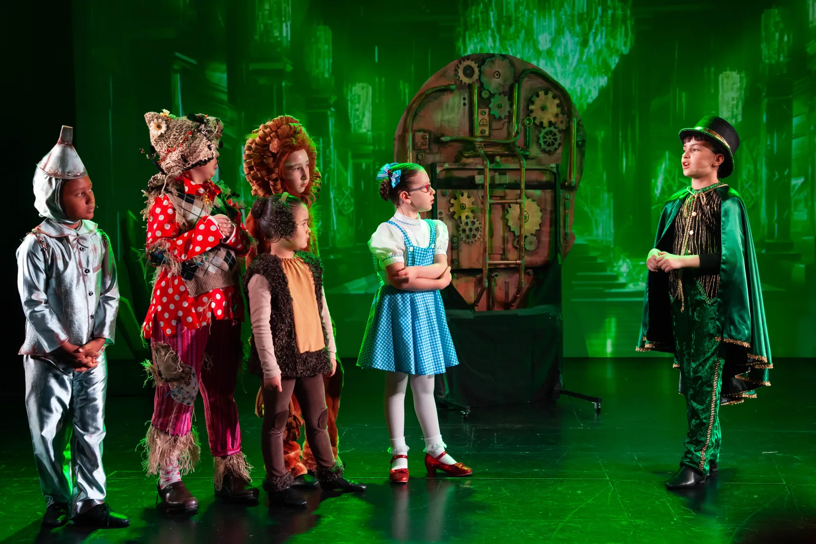 Starbright students performing The Wizard of Oz on the Starbright Theater stage, the Tin Man, Scarecrow, Lion, Toto, and Dorothy facing the Wizard under green light