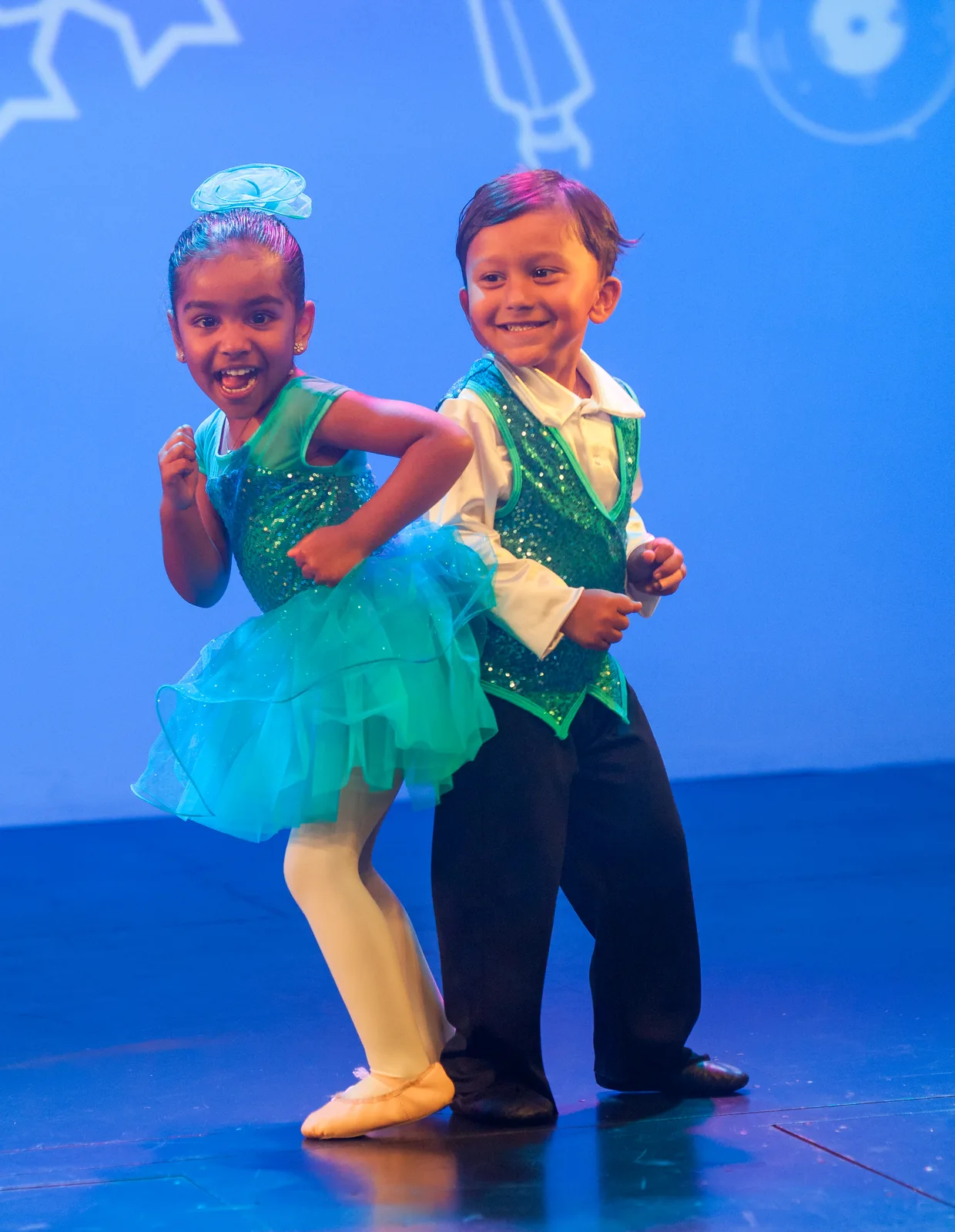 Two young Starbright dancers in sparkling teal costumes performing on the Starbright Theater stage