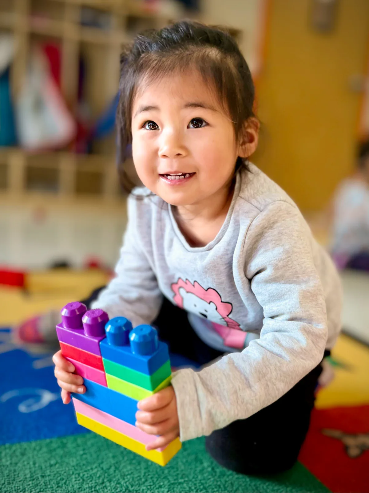 A smiling Starbright preschooler kneeling on the classroom rug, holding up a tower she has built from coloured blocks