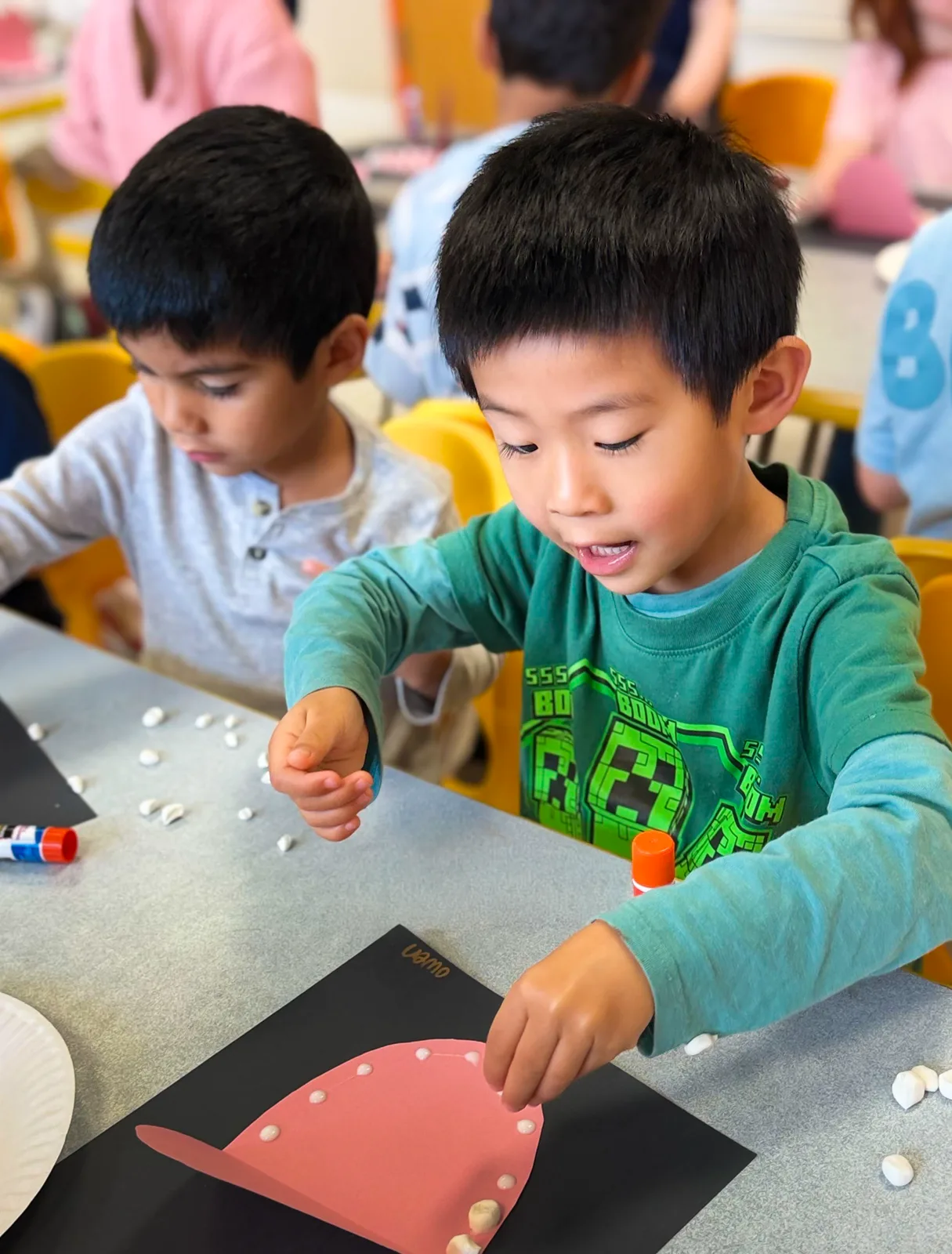 Two Starbright students at a classroom table, gluing small paper pieces onto a red paper shape