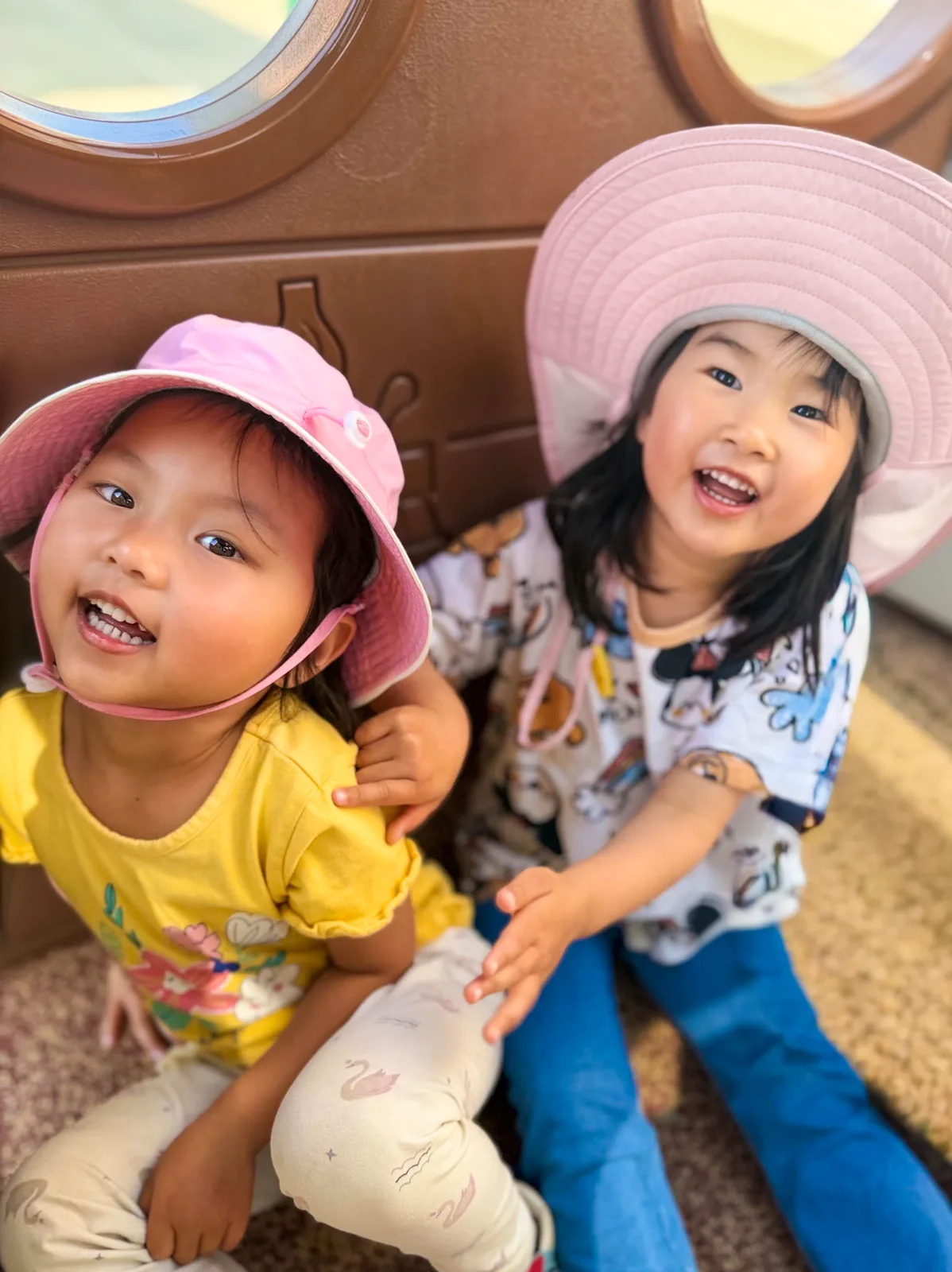 Two Starbright students in sun hats smiling together beside an outdoor playhouse